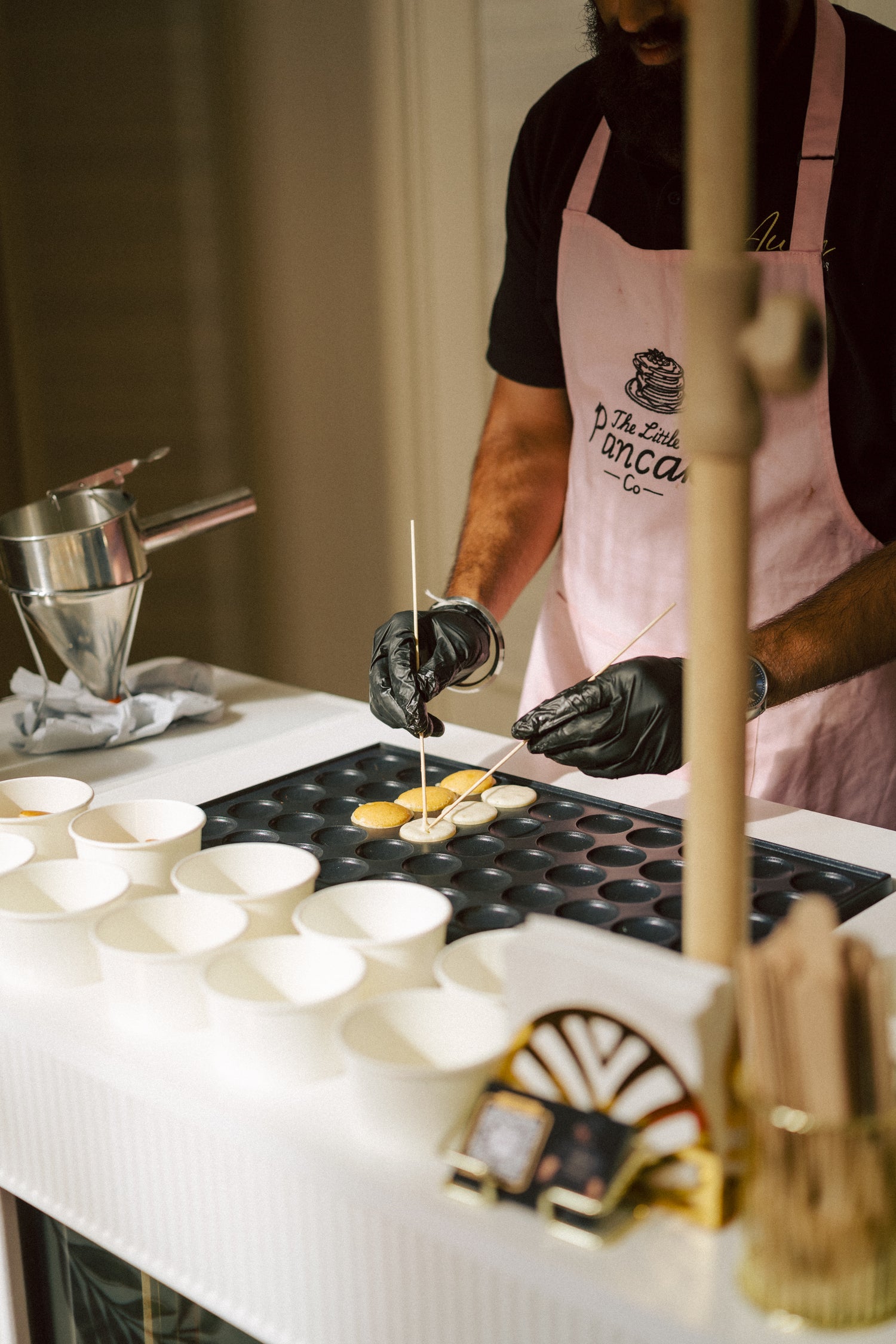 A chef wearing a pink apron with 'The Aura Events logo and black gloves is making mini pancakes on a white table with cart